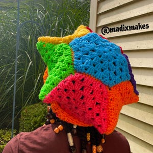 May include: A vibrant, multi-colored crochet hat with a patchwork design. The hat features bright hues of red, orange, yellow, green, blue, and purple. The text "@madixmakes" is visible on the side. The hat is worn by a person with dreadlocks.