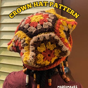 Op de afbeelding: Een handgemaakte gehaakte kroonhoed in bruin, geel en oranje tinten. De hoed heeft een textuur, geometrisch patroon en wordt gedragen door een persoon met dreadlocks versierd met houten kralen. De tekst "CROWN HAT PATTERN" wordt weergegeven.
