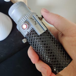 Può includere: Un accessorio per spada laser nero e argento con una luce rossa sul lato e un'impugnatura testurizzata. La spada laser è etichettata "NZ".