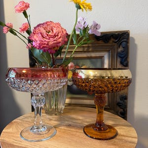 May include: Two vintage champagne coupe glasses with diamond-cut bowls. One glass has a pink rim, the other a gold rim. The glasses sit on a wooden table with a vase of flowers in the background.