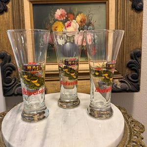 May include: Three clear Budweiser shot glasses with a gold rim and a colorful graphic of a building and water. The glasses are displayed on a white marble surface with a gold-colored decorative edge. A framed floral painting is in the background.