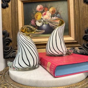 May include: Two glass duck figurines with white bodies and black vertical stripes. The ducks have gold-colored beaks and are displayed on a pink book with blue pages. The background includes a framed painting of flowers.