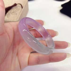May include: A translucent, lavender-colored jadeite bangle bracelet. The bracelet has a smooth, polished surface and a gradient effect, transitioning from a light purple to a clear, almost white color. The bracelet is held in a hand.