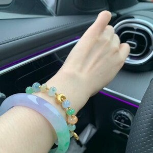 May include: Two jade bracelets. One is a light green and lavender bangle. The other is a gold chain bracelet with jade beads in shades of green, orange, and blue. The bracelets are on a person's wrist.