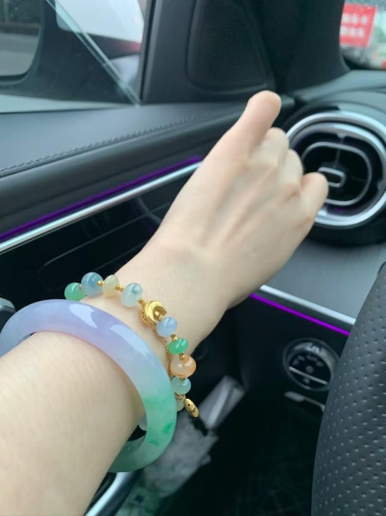 May include: Two jade bracelets. One is a light green and lavender bangle. The other is a gold chain bracelet with jade beads in shades of green, orange, and blue. The bracelets are on a person's wrist.