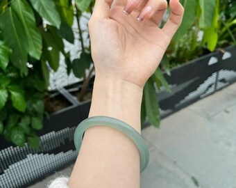 May include: A pale green jadeite bangle bracelet. The smooth, translucent bracelet is worn on the wrist. The background includes green foliage and a cream-coloured sweater.