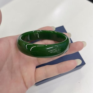 Può includere: Un braccialetto bangle in giada verde scuro lucido. Il braccialetto liscio e circolare è tenuto in mano, mostrando la sua superficie lucida. La giada è di un colore verde intenso e ricco, che riflette la luce. Un gioiello classico.