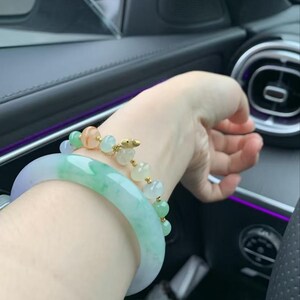 May include: A wrist adorned with two bracelets. One is a wide, translucent jade bangle with shades of green and lavender. The other is a delicate gold chain bracelet with colorful, round beads. The bracelets are displayed on a wrist.