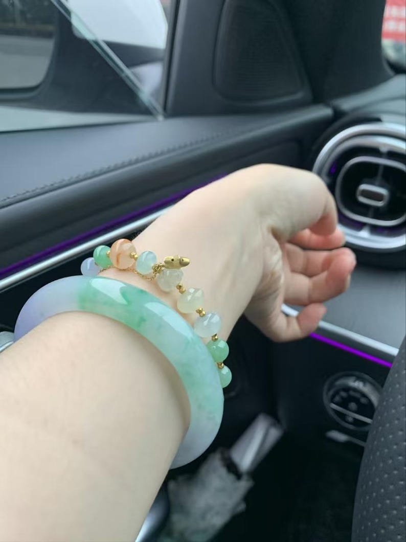 May include: A wrist adorned with two bracelets. One is a wide, translucent jade bangle with shades of green and lavender. The other is a delicate gold chain bracelet with colorful, round beads. The bracelets are displayed on a wrist.