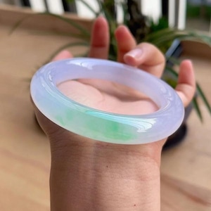 May include: A translucent jade bangle bracelet. The bracelet is a light lavender color with a section of vibrant green. The smooth, polished surface reflects light, highlighting the natural variations in the stone. This elegant piece is a classic jewelry item.
