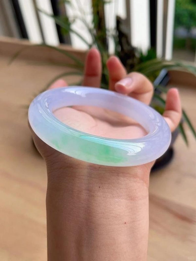 May include: A translucent jade bangle bracelet. The bracelet is a light lavender color with a section of vibrant green. The smooth, polished surface reflects light, highlighting the natural variations in the stone. This elegant piece is a classic jewelry item.