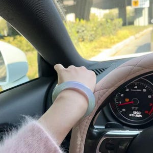 May include: A light green jade bangle bracelet worn on a person's wrist. The person is driving a car with a steering wheel and dashboard visible.