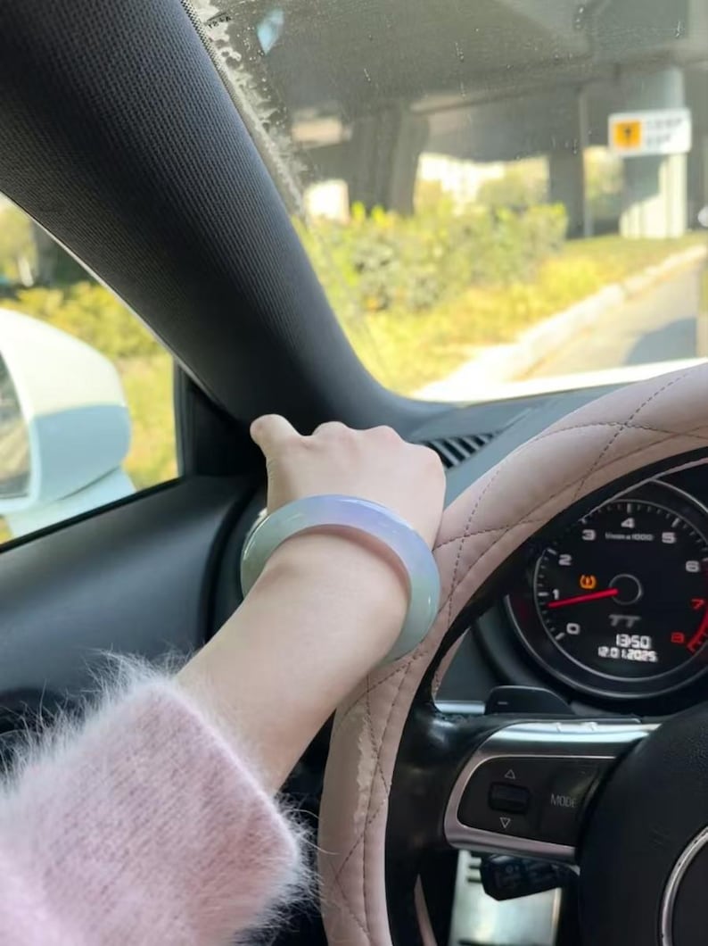 May include: A light green jade bangle bracelet worn on a person's wrist. The person is driving a car with a steering wheel and dashboard visible.