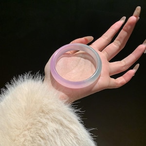 Può includere: Un braccialetto rotondo traslucido con una sfumatura di colori, che passa dal lavanda a un verde acqua pallido. Il braccialetto è tenuto contro uno sfondo scuro, mostrando la sua superficie liscia e lucida. Il design del braccialetto è semplice ed elegante.