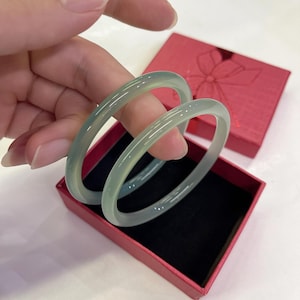 Pode incluir: Duas pulseiras de jade verde-claro são exibidas dentro de uma caixa de presente vermelha com um desenho de laço na tampa. As pulseiras são lisas e translúcidas, com uma sutil variação de cor natural. A caixa tem um interior preto.