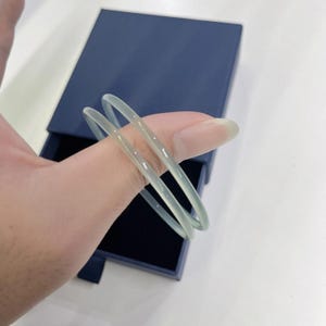 Op de afbeelding: Twee doorschijnende, lichtgroene armbanden worden door een hand vastgehouden, met een donkerblauwe doos op de achtergrond. De armbanden zijn dun en lijken gemaakt van een glad, gepolijst materiaal. De algehele esthetiek is minimalistisch en elegant.