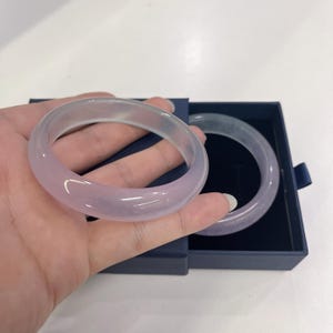 May include: Two translucent, light purple bangles. One is held in a hand, the other rests in a dark blue box. The bangles have a smooth, polished surface and a rounded shape. The color is a soft, pastel hue.