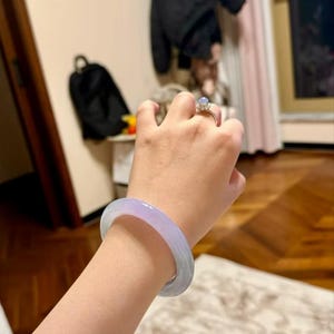 May include: A lavender jade bangle bracelet worn on a person's wrist. The bracelet is smooth and polished, with a light lavender colour. The person is wearing a ring with a clear stone on their finger.