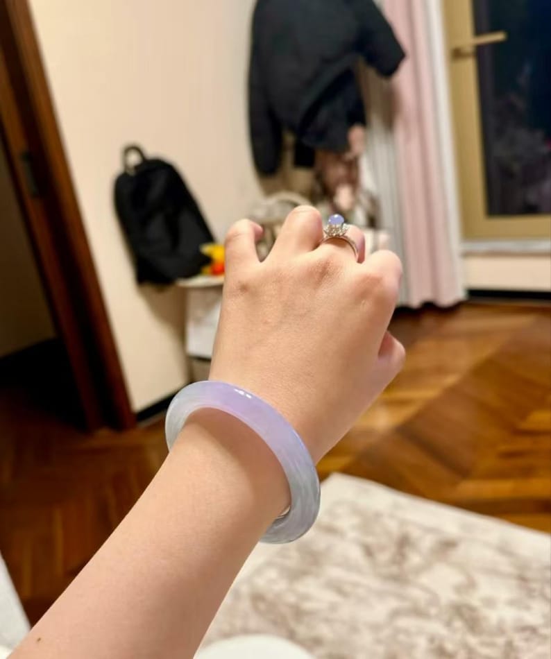 May include: A lavender jade bangle bracelet worn on a person's wrist. The bracelet is smooth and polished, with a light lavender colour. The person is wearing a ring with a clear stone on their finger.