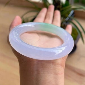 May include: A translucent jade bangle bracelet. The bracelet has a smooth, polished surface with a gradient of colors, transitioning from a pale lavender to a soft green hue. The bangle is held in a hand, showcasing its elegant design.