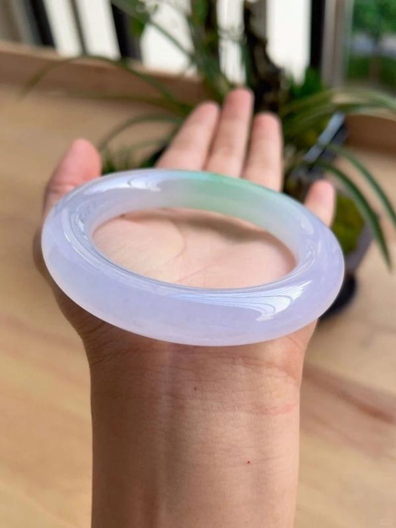 May include: A translucent jade bangle bracelet. The bracelet has a smooth, polished surface with a gradient of colors, transitioning from a pale lavender to a soft green hue. The bangle is held in a hand, showcasing its elegant design.