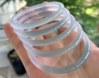 Peut inclure: Plusieurs bracelets en jade vert pâle translucide. Les bracelets circulaires et lisses sont empilés dans une main, mettant en valeur leur couleur délicate et leur finition polie. L'arrière-plan est flou, avec du feuillage vert.