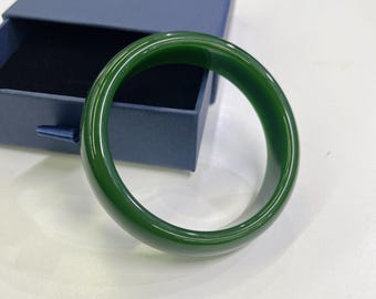 May include: A polished, jade-green bangle bracelet. The smooth, circular bracelet is displayed on a white surface next to an open, navy blue jewelry box. The bangle's color is a deep, rich green.