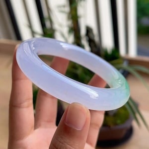 May include: A translucent jade bangle bracelet. The bracelet is a pale lavender color that transitions to a light green hue. The smooth, polished surface reflects light, showcasing the natural variations in the stone.