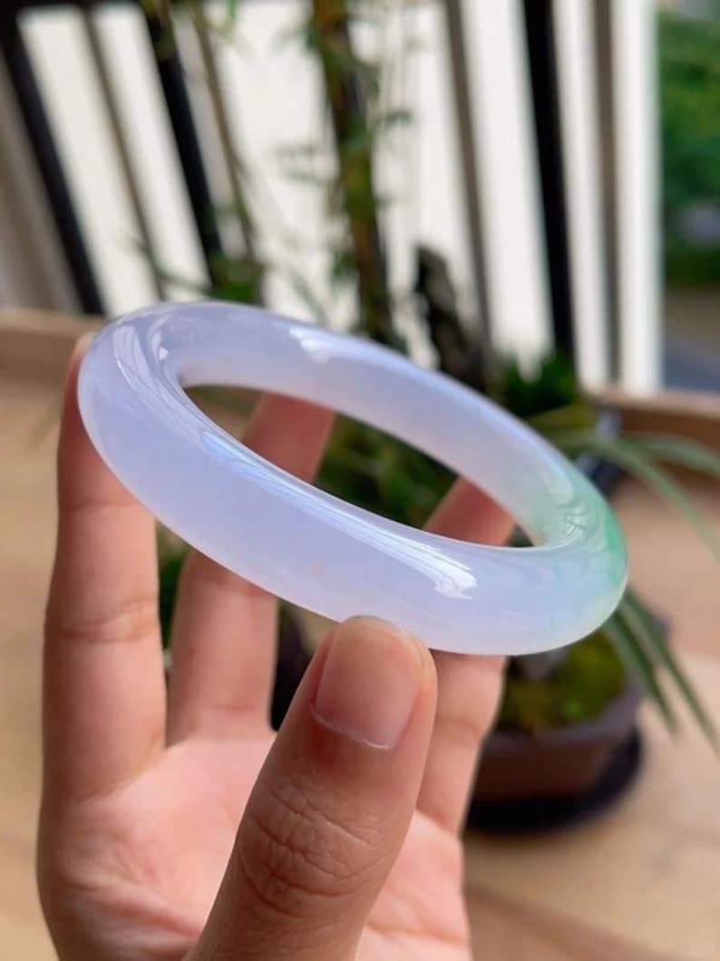 May include: A translucent jade bangle bracelet. The bracelet is a pale lavender color that transitions to a light green hue. The smooth, polished surface reflects light, showcasing the natural variations in the stone.