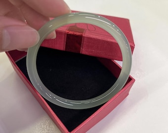 Könnte beinhalten: Ein hellgrünes Jade-Armreif-Armband. Das glatte, kreisförmige Armband wird über einer roten Geschenkbox mit schwarzem Innenraum gehalten. Der Jade hat eine durchscheinende Qualität, und die Box ist geöffnet.