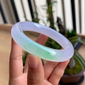 May include: A translucent jade bangle bracelet. The bracelet features a gradient of colors, transitioning from a pale lavender to a vibrant green. The polished surface reflects light, highlighting the natural variations in the stone.