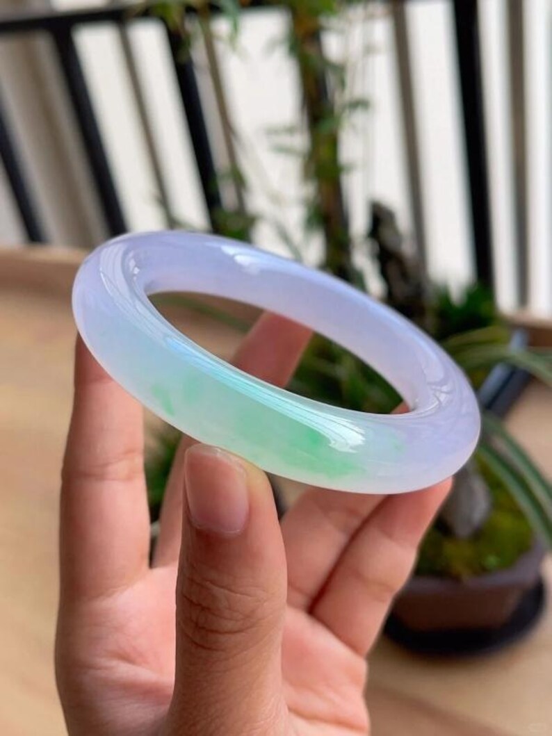 May include: A translucent jade bangle bracelet. The bracelet features a gradient of colors, transitioning from a pale lavender to a vibrant green. The polished surface reflects light, highlighting the natural variations in the stone.