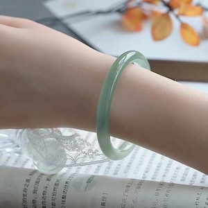 Könnte beinhalten: Ein hellgrünes Jade-Armreif-Armband. Die glatte, polierte Oberfläche reflektiert das Licht und zeigt die natürlichen Farbvariationen. Das Armband hat ein klassisches, kreisförmiges Design, das für den Alltag geeignet ist. Der Jade ist ein blasses, durchscheinendes Grün.