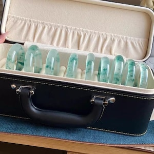 Könnte beinhalten: Eine schwarze Ledertasche mit Griff, die mit weißem Samt ausgekleidet ist und eine Sammlung von jadegrünen Armreifen enthält.