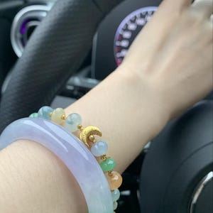 May include: A wrist adorned with two bracelets. One is a wide, translucent jade bangle with green and lavender hues. The other is a beaded bracelet with a gold crescent moon charm and colorful beads in shades of green, blue, and orange.