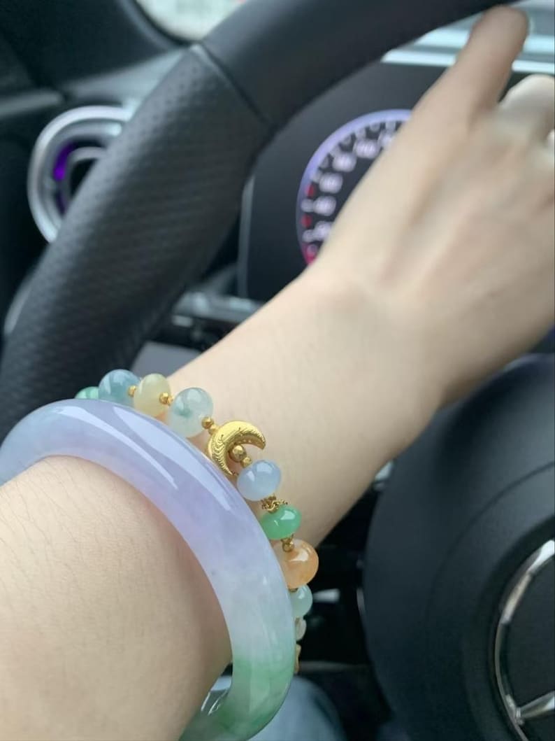 May include: A wrist adorned with two bracelets. One is a wide, translucent jade bangle with green and lavender hues. The other is a beaded bracelet with a gold crescent moon charm and colorful beads in shades of green, blue, and orange.