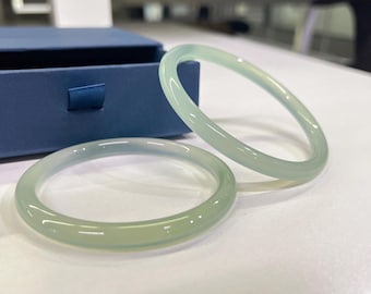 May include: Two pale green jadeite bangles sit on a white surface, near a blue box. The smooth, translucent bracelets have a rounded shape. The lighting highlights their subtle colour variations and polished finish.