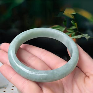 May include: A light green jade bangle bracelet. The smooth, polished surface of the bracelet reflects light, showcasing its natural color variations. The jade is a pale green hue, with subtle variations in tone. The bracelet is a circular shape.