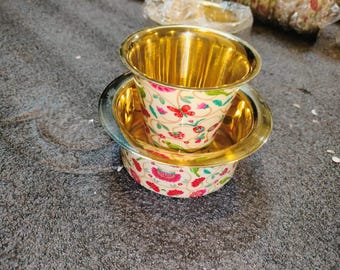May include: A decorative tea cup and saucer set. The interior of the cup and saucer are gold-coloured. The exterior is cream-coloured with a floral pattern in red, pink, and green.