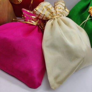 Może przedstawiać: Zbliżenie na kilka kolorowych, materiałowych woreczków. Woreczki są w odcieniach różu, kremu i zieleni. Każdy woreczek jest u góry zawiązany złotą wstążką. Tkanina ma delikatny połysk.