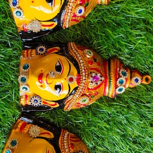 May include: Three decorative face masks with yellow skin, black hair, and elaborate gold and jewel-toned headdresses. The masks are arranged on green grass, showcasing intricate details and vibrant colours.