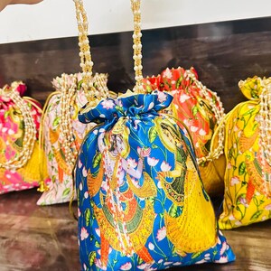 Pode incluir: Uma bolsa de cordão azul com uma alça de pérolas douradas e brancas. A bolsa apresenta uma ilustração colorida de um casal com roupas tradicionais indianas. A bolsa é decorada com guarnições douradas e um padrão floral.