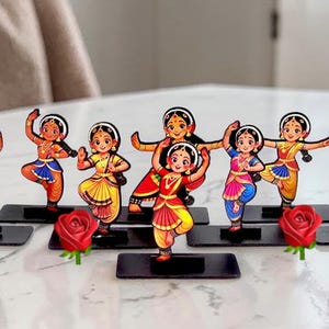 May include: A collection of nine individual flat acrylic cutouts featuring cartoon-style dancers in traditional Indian attire, each mounted on a small black rectangular base. The dancers wear colorful outfits in shades of red, blue, yellow, and green.