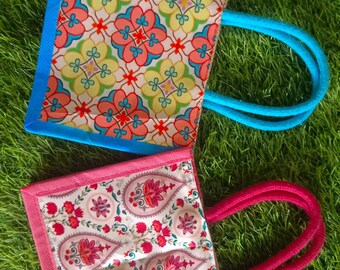 May include: Two small tote bags with colorful floral patterns and rope handles. One bag has a blue border and handle, the other has a pink border and handle. The bags are on a green grassy surface.