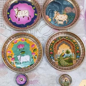 Pode incluir: Quatro pratos decorativos de metal com desenhos pintados intrincados. Cada prato apresenta uma cena diferente: uma vaca, uma vaca com um bezerro, uma vaca com flores de lótus e um pavão. Os pratos têm uma borda dourada e estão sobre uma superfície estampada.