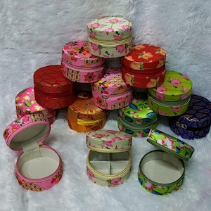 Puede incluir: Surtido de cajas de joyería redondas de tela de colores vivos con motivos florales y geométricos. Las cajas están apiladas y algunas están abiertas para revelar un interior blanco suave.