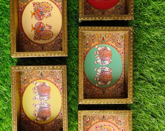 May include: Five decorative framed art pieces with gold-coloured frames. Each frame contains a circular design with colourful illustrations of birds and elephants on various coloured backgrounds, including red, green, and yellow. The frames are arranged on green grass.