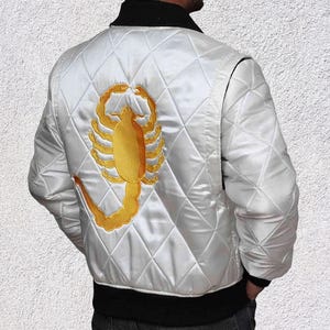 May include: White satin bomber jacket with a quilted pattern, black collar and trim. The back features a large, embroidered gold scorpion design. A classic jacket style, suitable for various occasions.