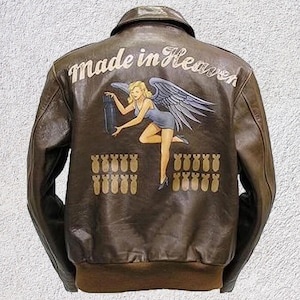 Può includere: Giacca in pelle marrone in stile bomber. Il retro presenta un'immagine dipinta di una donna con ali d'angelo che tiene una bomba, con la scritta "Made in Heaven" sopra. Sotto l'immagine ci sono diverse bombe dipinte.