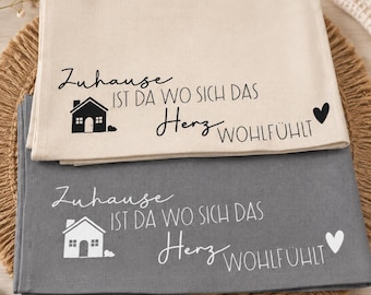 Könnte beinhalten: Zwei rechteckige Küchentücher, eines in Beige und eines in Dunkelgrau, jeweils mit einer Grafik eines Hauses, eines Herzens und dem deutschen Text "Zuhause ist da wo sich das Herz wohlfühlt" in einer Kombination aus Schreib- und Druckschrift.